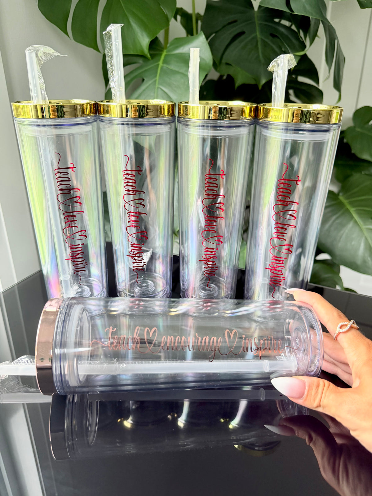 Five clear double-wall insulated tumblers with gold lids and clear straws are displayed on a glossy black surface, with a hand holding one tumbler in the foreground. The tumblers have red script text reading 'teach love inspire'.