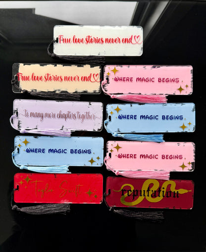 A set of assorted acrylics handmade bookmarks with tassel ends, shown in multiple colors on a dark reflective surface. Each piece has printed text such as 'True love stories never end', 'Where magic begins', and 'Reputation', Taylor Swift, true love stories never end, to many more chapters together.