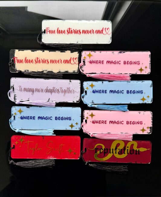 A set of assorted acrylics handmade bookmarks with tassel ends, shown in multiple colors on a dark reflective surface. Each piece has printed text such as 'True love stories never end', 'Where magic begins', and 'Reputation', Taylor Swift, true love stories never end, to many more chapters together.