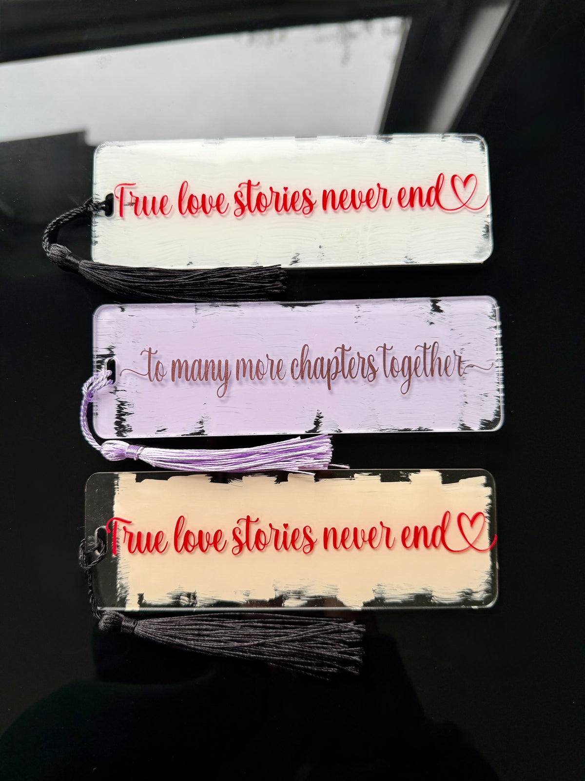 Three rectangular book bookmarks with tassel ties are shown on a dark reflective surface. The top bookmark is white with red text reading 'True love stories never end' and a small red heart.