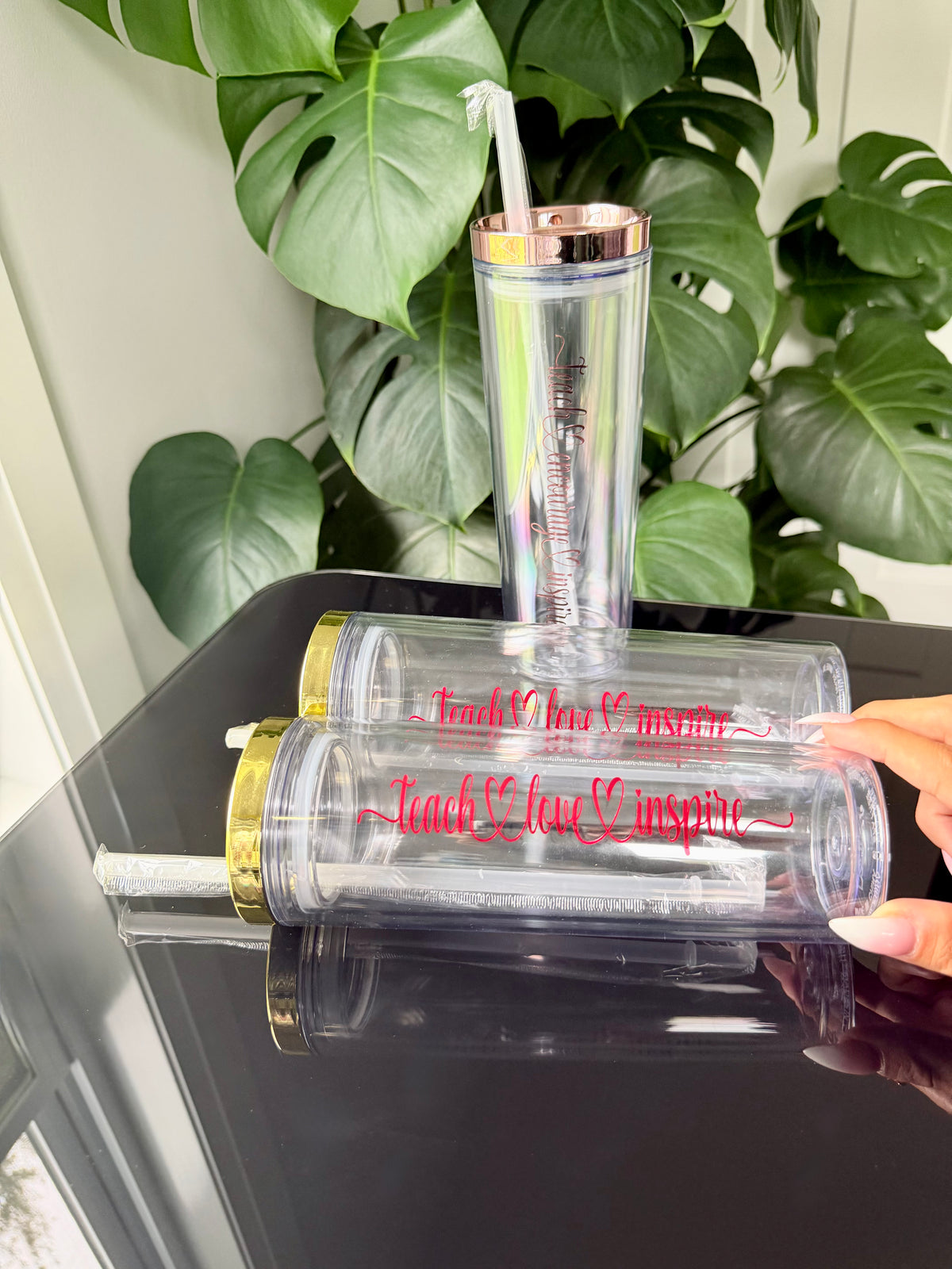 Two clear plastic tumblers with gold and rose gold lids are displayed on a black reflective surface, with a hand holding one tumbler. 