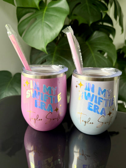 Two insulated stemless tumblers with clear lids and pink straws, displayed on a reflective dark surface with green plant leaves in the background. One tumbler is pink with blue and gold text reading 'IN MY SWIFTIE ERA Taylor Swift,' and the other is opal with blue and gold text reading 'IN MY SWIFTIE ERA Taylor Swift.