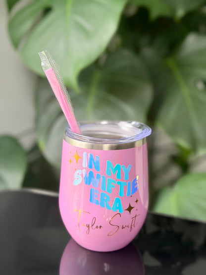 A pink insulated stemless tumbler with a clear lid and pink straw, featuring the text 'IN MY SWIFTIE ERA Taylor Swift' in blue and gold lettering.