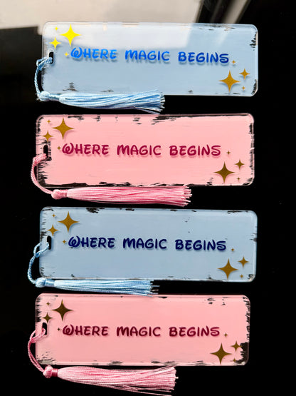 Four rectangular acrylic bookmarks with tassel ties are arranged vertically on a dark background. Two are light blue and two are pink, each printed with the phrase “WHERE MAGIC BEGINS.