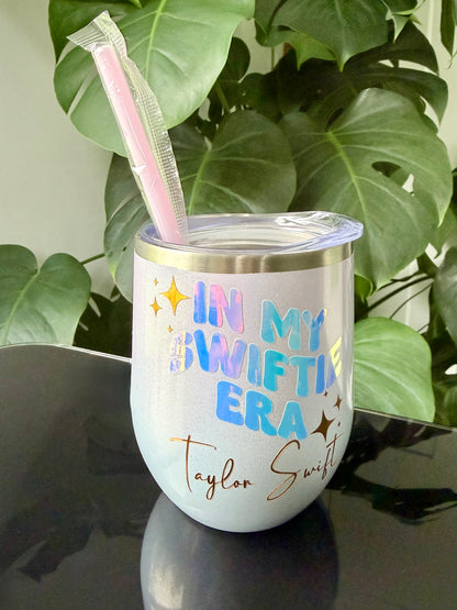 A opal insulated tumbler with a clear lid and pink straw, printed with the phrase 'IN MY SWIFTIE ERA Taylor Swift' in pastel colors and gold lettering. 